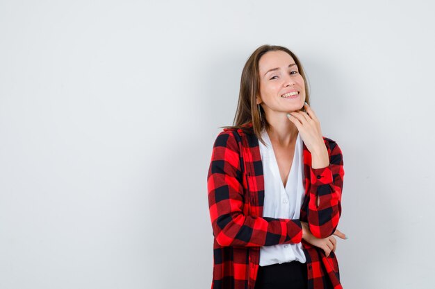 Jonge mooie vrouw die zich voordeed terwijl ze met de hand onder de kin in een casual outfit zit en er zalig uitziet, vooraanzicht.