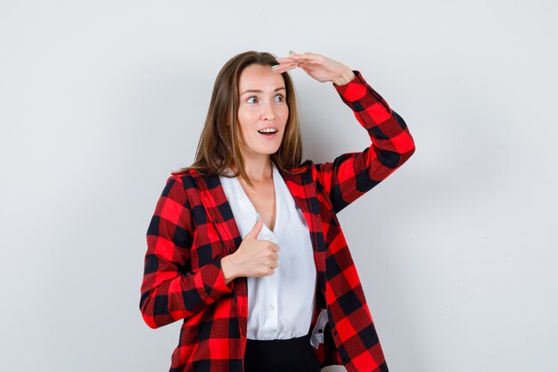 Jonge mooie vrouw duim opdagen, met de hand boven het hoofd in casual outfit en verbaasd kijken, vooraanzicht.