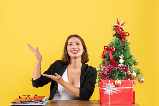 Jonge mooie vrouw iets trots tonen aan een tafel in de buurt van versierde kerstboom op kantoor op geel