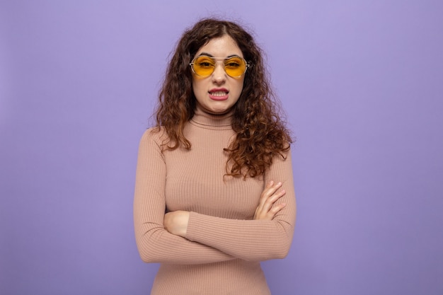 Jonge mooie vrouw in beige coltrui met een gele bril die er geïrriteerd en geïrriteerd uitziet met gekruiste armen