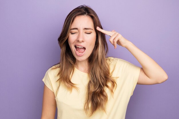 Jonge mooie vrouw in beige t-shirt die verward en verrast kijkt en een pistoolgebaar maakt over de tempel die over paars staat