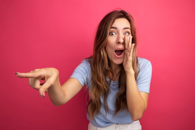 Jonge mooie vrouw in blauw t-shirt die een geheim vertelt met de hand in de buurt van de mond wijzend met de wijsvinger naar iets dat over een roze achtergrond staat