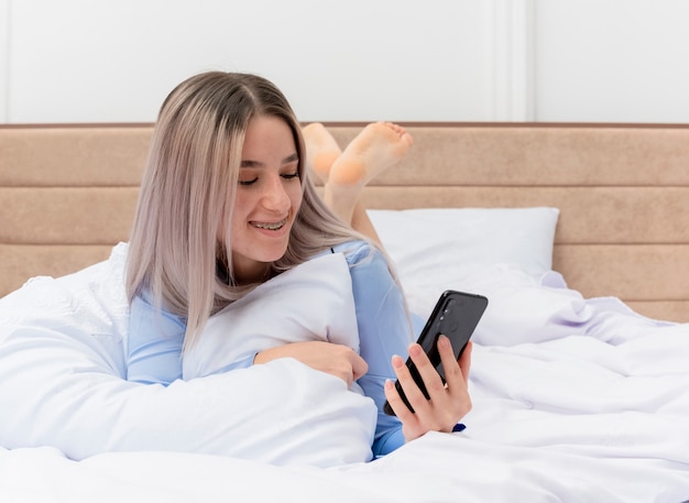 Jonge mooie vrouw in blauwe pyjama's die op bed ligt met behulp van smartphone gelukkig en positief rustend in slaapkamerinterieur