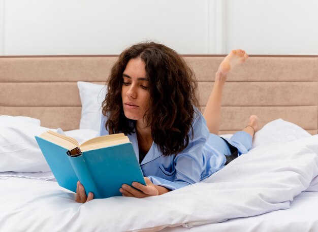 Jonge mooie vrouw in blauwe pyjama's liggend op bed leesboek in slaapkamer interieur op lichte achtergrond