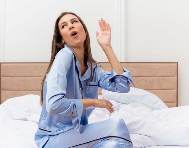 Jonge mooie vrouw in blauwe pyjama zittend op bed hapy en opgewonden op zoek vreugdevol in slaapkamer interieur op lichte achtergrond