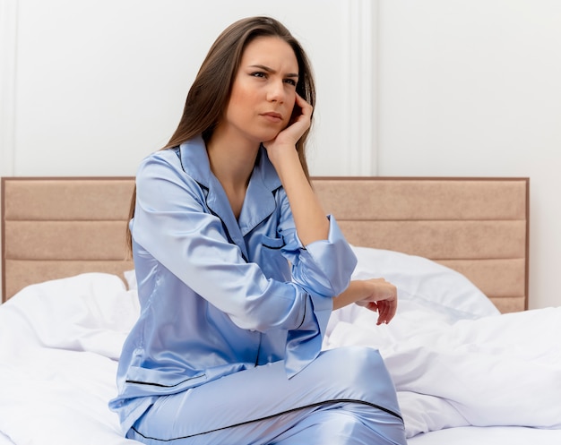 Jonge mooie vrouw in blauwe pyjama zittend op bed lookign opzij met ernstige fronsend gezicht in slaapkamer interieur op lichte achtergrond