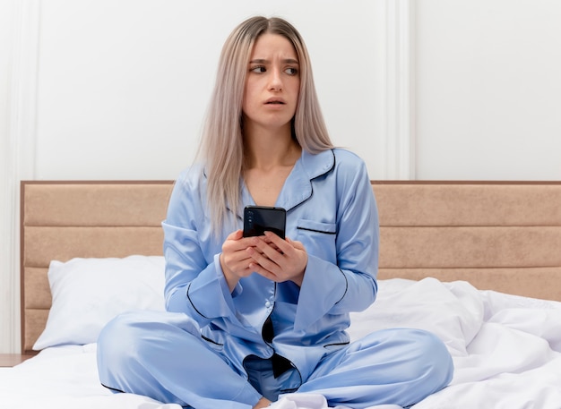 Jonge mooie vrouw in blauwe pyjama zittend op bed met smartphone op zoek opzij verward in slaapkamer interieur op lichte achtergrond