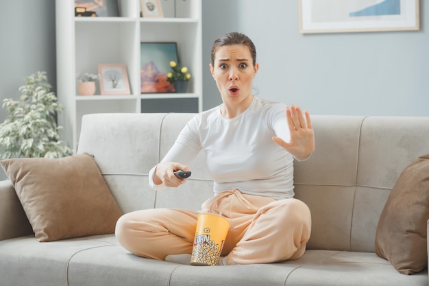 jonge mooie vrouw in casual kleding zittend op een bank thuis interieur met emmer popcorn met afstandsbediening kijken naar camera bezorgd zijn stop gebaar met hand maken