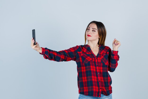 Jonge mooie vrouw in casual shirt die selfie neemt, winnaargebaar toont en er trots uitziet, vooraanzicht.