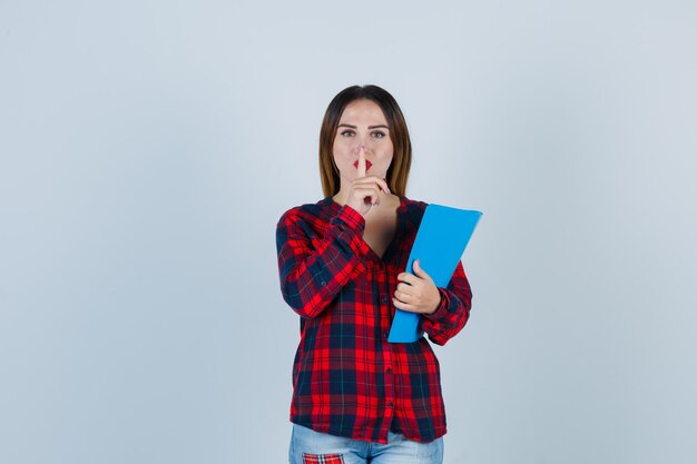 Jonge mooie vrouw in casual shirt met map, stiltegebaar tonend en gefocust, vooraanzicht.