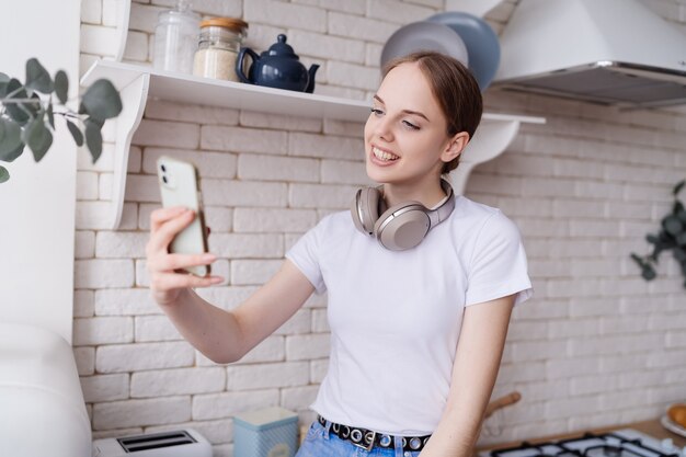 Jonge, mooie vrouw in casual zit op de keukentafel met een koptelefoon en maakt videocall