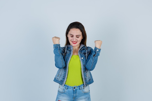 Jonge mooie vrouw in denim outfit die winnaargebaar toont en er trots uitziet, vooraanzicht.