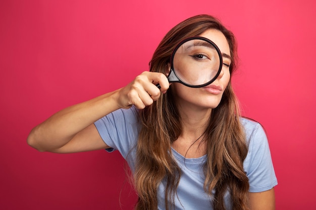 Jonge, mooie vrouw in een blauw t-shirt die door een vergrootglas naar de camera kijkt met belangstelling voor roze