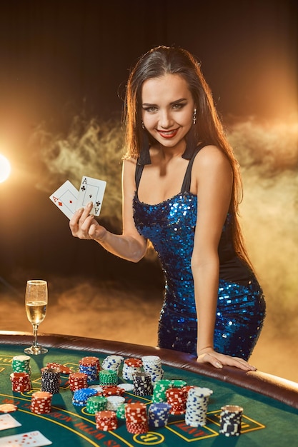Jonge mooie vrouw in een blauwe glanzende jurk poseert in de buurt van pokertafel in luxe casino. Vrouw speler. Passie, kaarten, chips, alcohol, dobbelstenen, gokken, casino - het is als vrouwelijk amusement. Gevaarlijk plezier