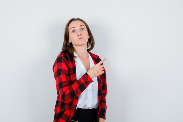 Jonge, mooie vrouw in een casual outfit die naar de rechterbovenhoek wijst en er bang uitziet, vooraanzicht.