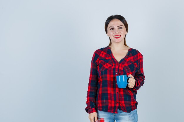 Jonge, mooie vrouw in een casual shirt, jeans die naar voren kijkt terwijl ze een kopje vasthoudt en er vrolijk uitziet, vooraanzicht.