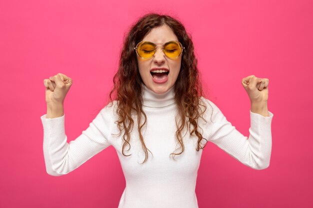 Jonge, mooie vrouw in een witte coltrui met een gele bril die vuisten opheft en schreeuwt met een geïrriteerde uitdrukking die op roze staat