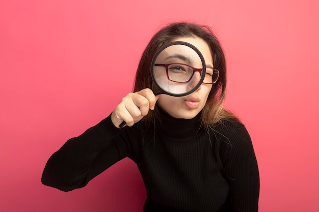 Gratis foto jonge mooie vrouw in een zwarte coltrui en glazen die voorzijde door vergrootglas bekijken die zich over roze muur bevinden