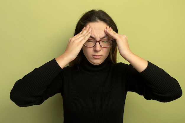 Jonge mooie vrouw in een zwarte coltrui en glazen kijken naar de voorkant aanraken van haar tempels die lijden aan hoofdpijn die zich over lichte muur bevindt