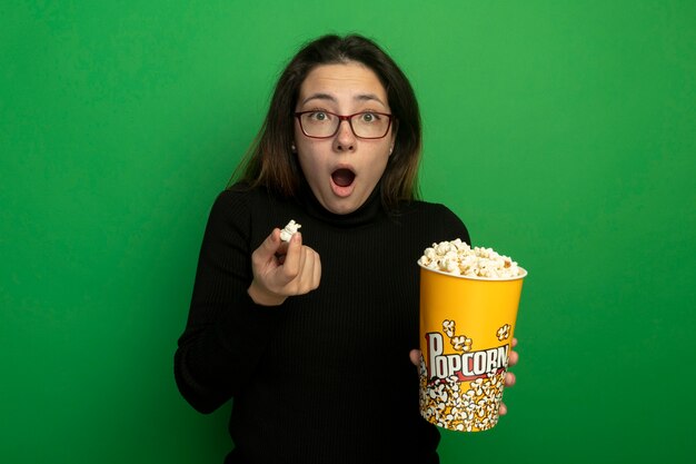 Jonge mooie vrouw in een zwarte coltrui en glazen met emmer met popcorn kijken naar voorkant verbaasd en verrast staande over groene muur