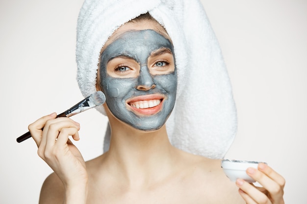 Jonge mooie vrouw in handdoek op hoofd die gezicht behandelen met masker het glimlachen. Schoonheid cosmetologie en spa.