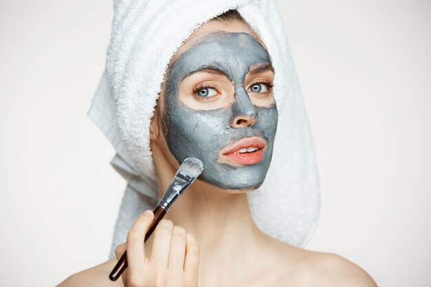 Jonge mooie vrouw in handdoek op hoofd die gezicht behandelen met masker het glimlachen. Schoonheid cosmetologie en spa.
