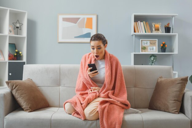 Jonge mooie vrouw in huiskleren zittend op een bank onder deken met smartphone die er verbaasd en geschokt uitziet om tijd thuis door te brengen Gratis Foto