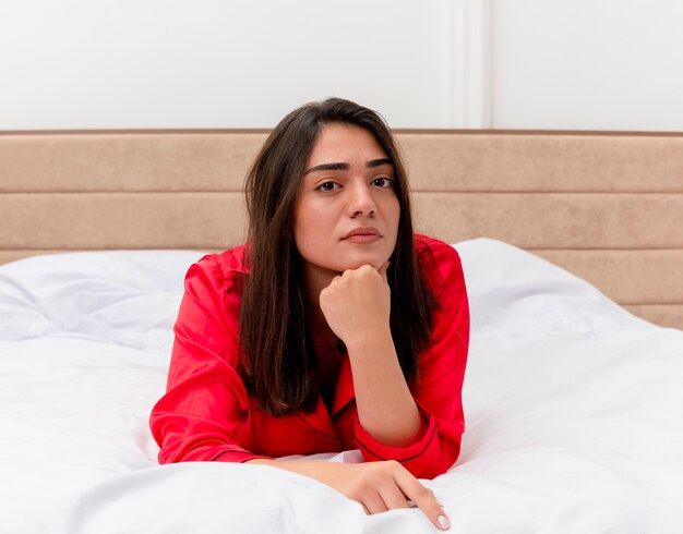 Jonge mooie vrouw in rode pyjama's liggend op bed camera kijken met peinzende uitdrukking op gezicht denken in interieur op lichte achtergrond