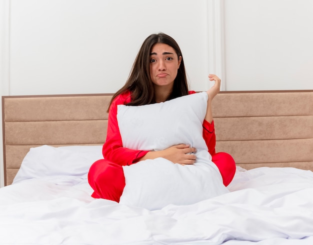 Jonge mooie vrouw in rode pyjama zittend in bed met kussen met droevige uitdrukking in slaapkamer interieur