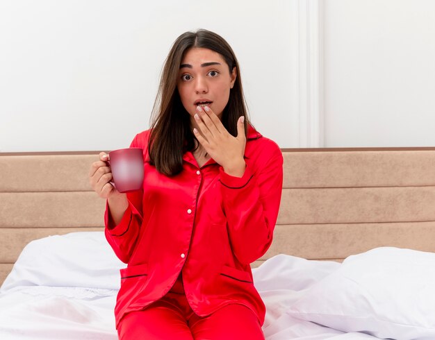 Jonge mooie vrouw in rode pyjama zittend op bed met kopje koffie kijken camera wordt verrast in slaapkamer interieur op lichte achtergrond