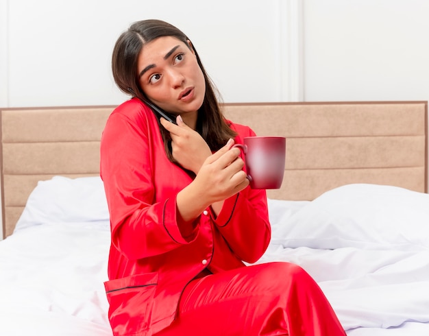 Jonge mooie vrouw in rode pyjama zittend op bed met kopje koffie praten op mobiele telefoon op zoek verward in slaapkamer interieur op lichte achtergrond