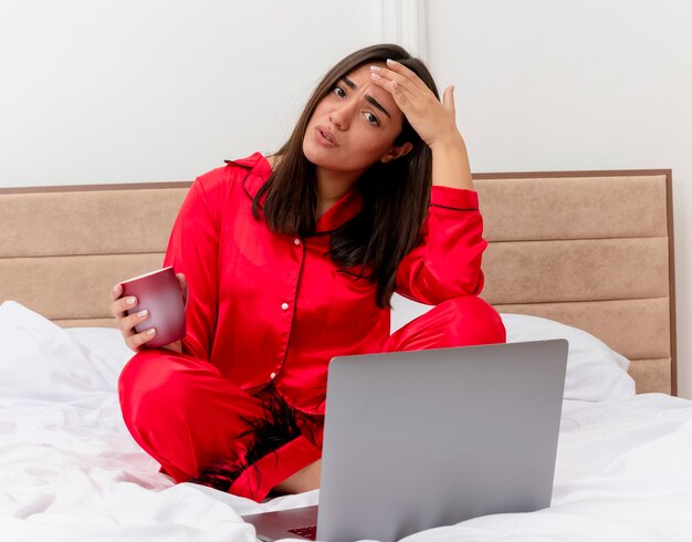 Jonge mooie vrouw in rode pyjama zittend op bed met laptop en kopje koffie kijken camera wordt verward en ontevreden in slaapkamer interieur op lichte achtergrond