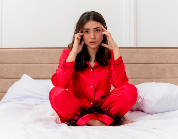 Jonge mooie vrouw in rode pyjama zittend op bed op zoek moe en verveeld aanraken van haar tempels in slaapkamer interieur op lichte achtergrond