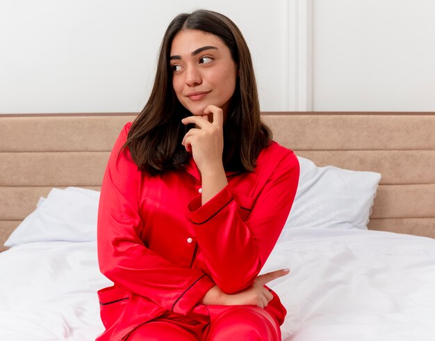 Jonge mooie vrouw in rode pyjama zittend op bed opzij kijken met peinzende uitdrukking denken en glimlachen in slaapkamer interieur op lichte achtergrond