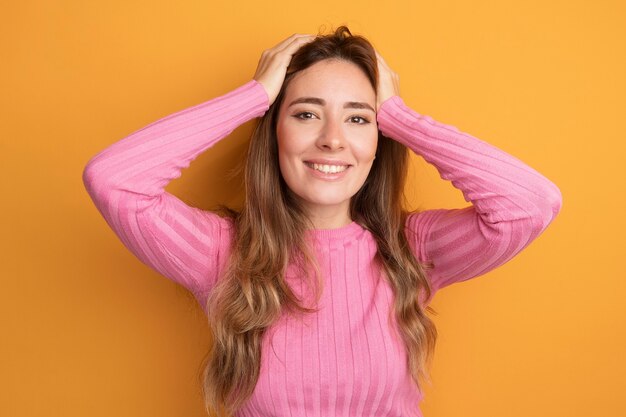 Jonge, mooie vrouw in roze top die blij en opgewonden naar de camera kijkt met de handen op haar hoofd over een oranje achtergrond