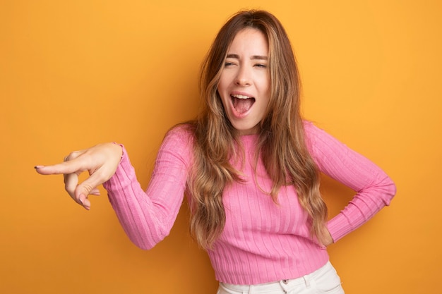 Jonge, mooie vrouw in roze top die opzij kijkt, gelukkig en vrolijk, wijzend met de wijsvinger naar iets