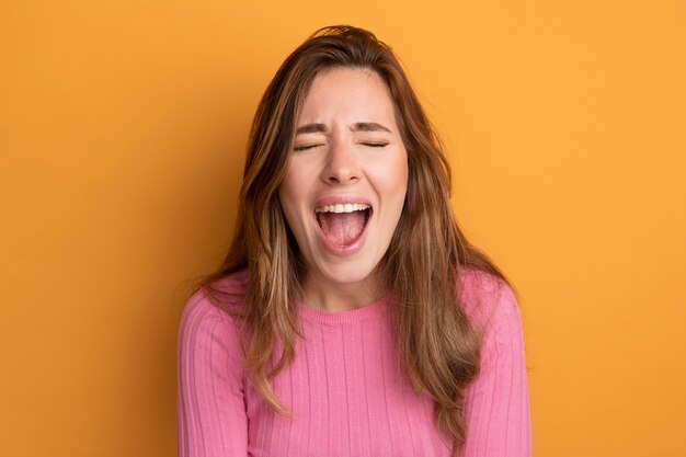 Jonge mooie vrouw in roze top, gek blij en opgewonden schreeuwend over oranje achtergrond