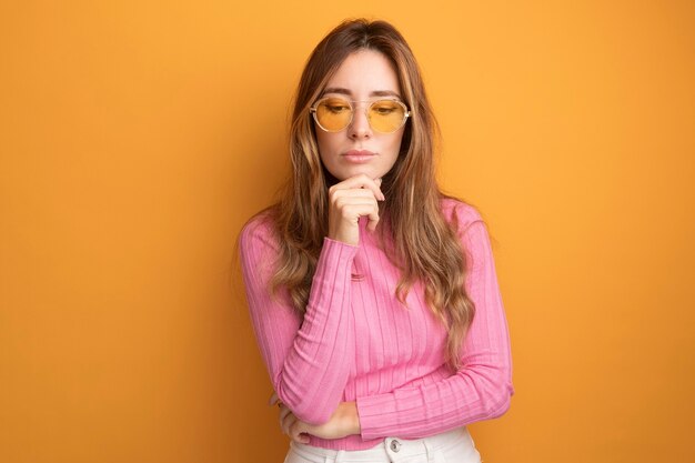 Jonge mooie vrouw in roze top met een bril die neerkijkt met de hand op de kin met een peinzende uitdrukking die over een oranje achtergrond denkt