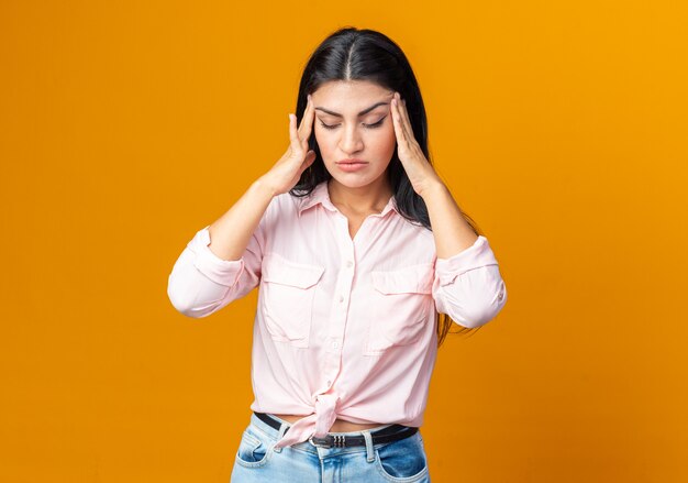 Jonge mooie vrouw in vrijetijdskleding die er onwel uitziet en haar slapen aanraakt die lijdt aan hoofdpijn die over een oranje muur staat