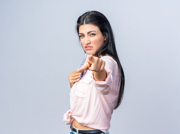 Jonge, mooie vrouw in vrijetijdskleding die met de wijsvinger wijst en kijkt met een boos gezicht dat over een witte muur staat