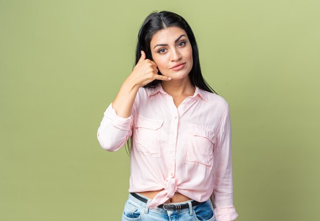 Jonge, mooie vrouw in vrijetijdskleding die naar de voorkant kijkt en zelfverzekerd glimlacht en me een gebaar maakt dat over een groene muur staat