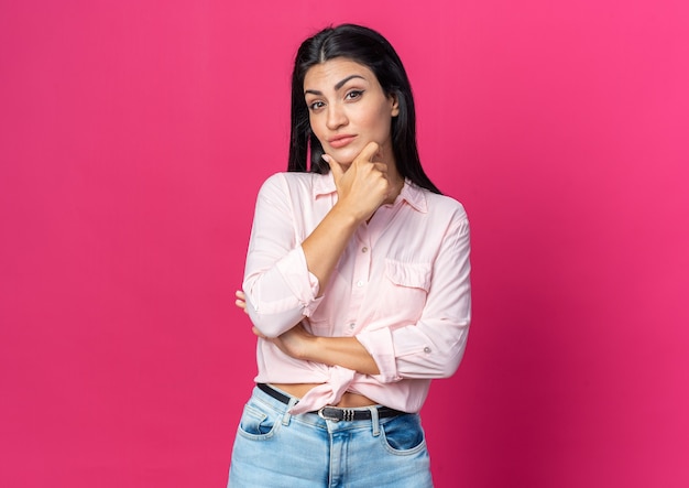 Jonge, mooie vrouw in vrijetijdskleding die naar de voorkant kijkt met de hand op de kin die over een roze muur staat te denken