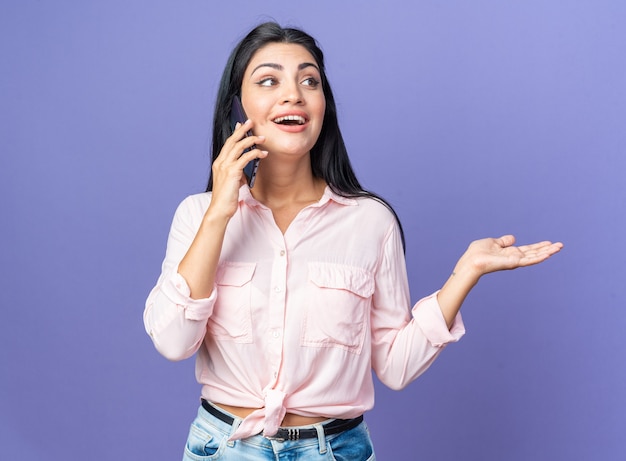 Jonge mooie vrouw in vrijetijdskleding die vrolijk gelukkig en positief glimlacht terwijl ze op een mobiele telefoon praat die over een blauwe muur staat