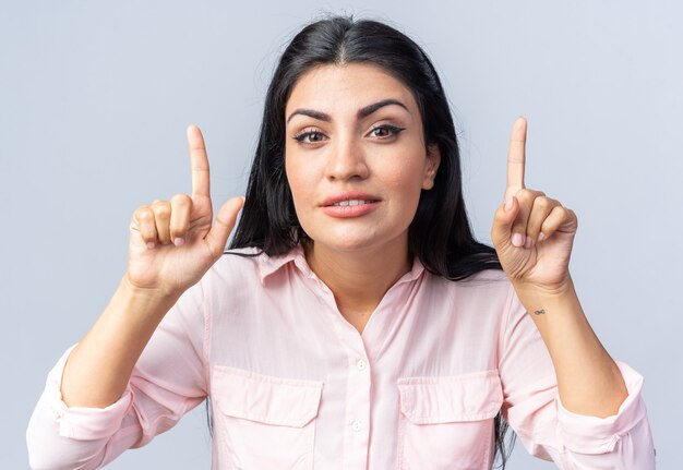 Jonge mooie vrouw in vrijetijdskleding glimlachend zelfverzekerd wijzend met wijsvingers omhoog staande over een witte muur