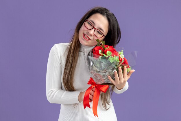 jonge mooie vrouw in vrijetijdskleding met boeket rode rozen gelukkig en positief glimlachen gevoel dankbaar Valentijnsdag concept staande over paarse muur