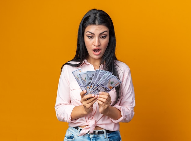 Jonge mooie vrouw in vrijetijdskleding met contant geld kijkend naar geld verbaasd en verrast Gratis Foto