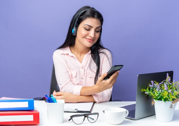 Jonge mooie vrouw in vrijetijdskleding met een hoofdtelefoon die een smartphone vasthoudt en ernaar kijkt glimlachend zittend aan de tafel met een laptop over een blauwe muur die op kantoor werkt