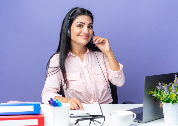 Jonge mooie vrouw in vrijetijdskleding met een hoofdtelefoon met microfoon die er zelfverzekerd uitziet glimlachend zittend aan de tafel met laptop over blauwe achtergrond die op kantoor werkt