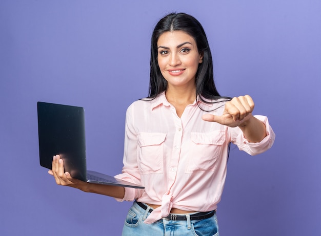 Jonge, mooie vrouw in vrijetijdskleding met een laptop die er glimlachend uitziet en met de duim naar de laptop wijst