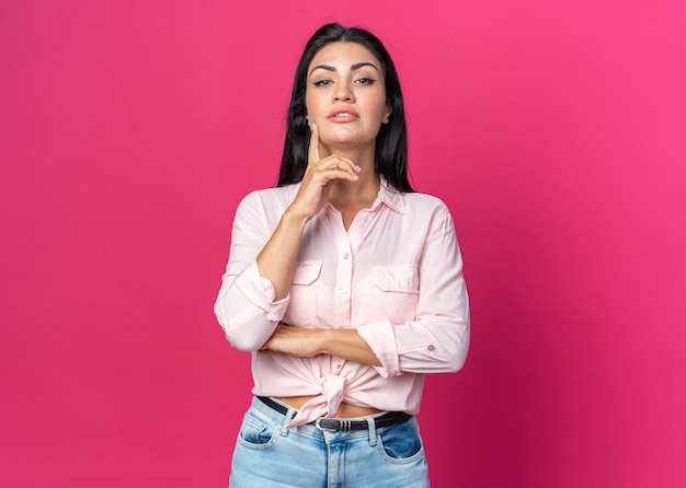 Jonge, mooie vrouw in vrijetijdskleding met een serieuze zelfverzekerde uitdrukking met een glimlach op een slim gezicht dat over een roze muur staat